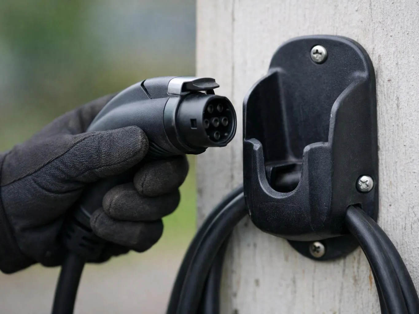 A gloved hand holds an electric vehicle charging plug near a wall-mounted charging holster outdoors